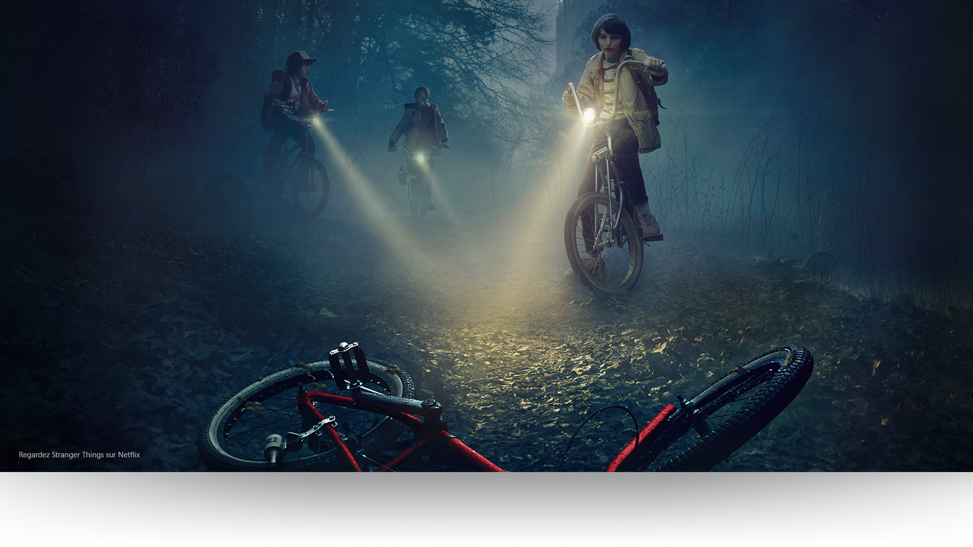 Stranger Things - Dustin, Lucas et Mike éclairent un vélo abandonné sur un sentier forestier lugubre.